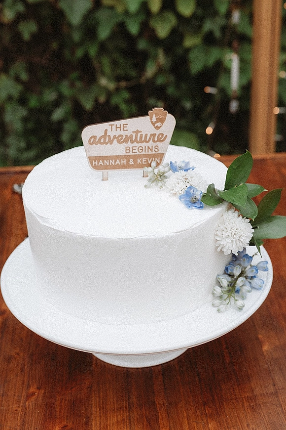 Wedding cake with a simple white buttercream finish, floral cluster and greenery, topped with a wooden topper on a pedestal amid string lights