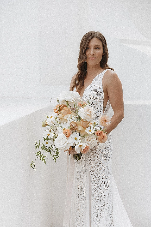 Bridal portrait of a bride holding bouquet with peach and white roses, daisies, and greenery in a lace deep V dress against a white studio backdrop