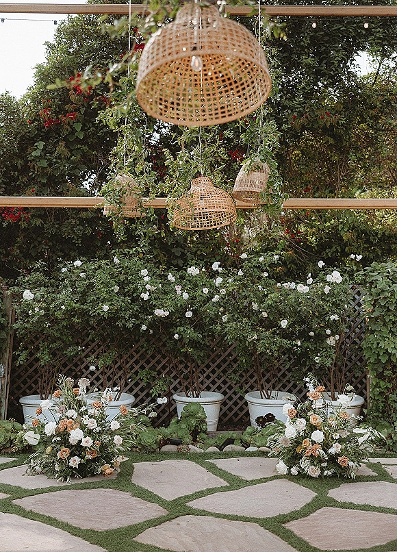 Ceremony backdrop decor with hanging wicker lanterns, lush greenery, and string lights above rose ground florals by a garden lattice fence