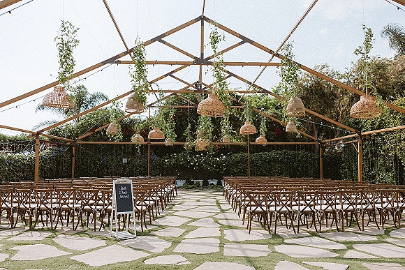 Ceremony setup under a clear tent wedding ceremony with wooden chairs, hanging greenery, and woven pendant lights along a stone paver aisle in a garden