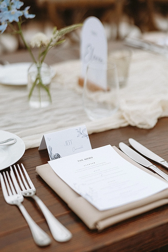 Wedding place setting with a wedding menu card on a neutral linen napkin, silver flatware, white plate, and bud vase on a wood reception table