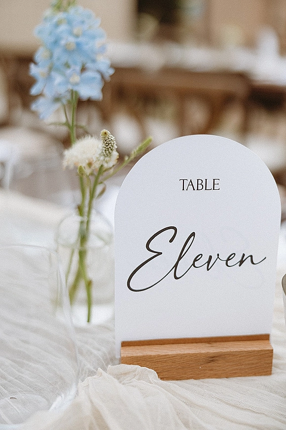 Wedding table number card in a wooden holder beside a bud vase of blue and white flowers on a gauze runner at reception tables