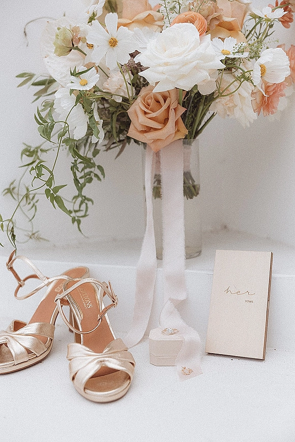 Wedding details flat lay with bridal details flatlay bouquet of peach roses and white cosmos, vow book, ring box, earrings and heels on white tabletop