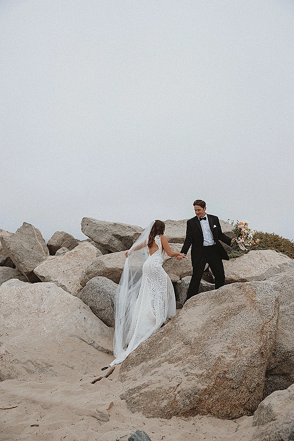 Couple portrait of bride and groom holding hands on a rocky shoreline, bride in wedding dress with long veil in the wind under cloudy sky