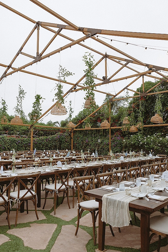 Outdoor reception setup with long banquet tables wedding, cross back chairs, and neutral linens under a pergola with rattan lights and greenery