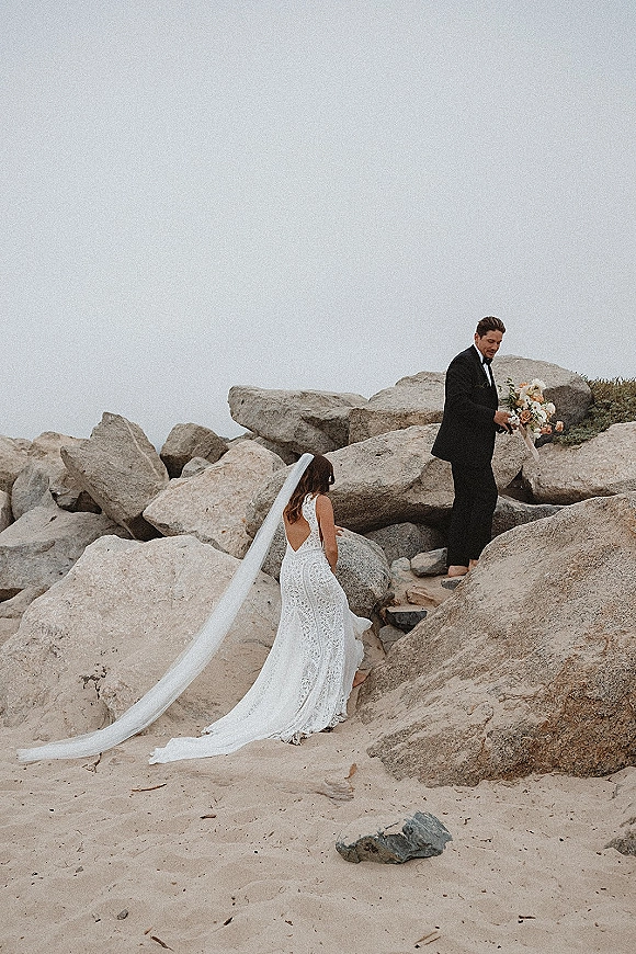 Couple portrait of bride in lace wedding dress with long veil and bouquet beside groom in black suit on a rocky beach under overcast sky