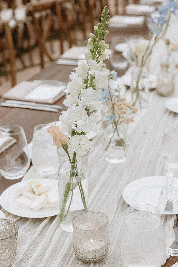 Reception tablescape with wedding bud vase centerpiece of white flowers and a peach rose, glass votive candles, and airy place settings on a long table