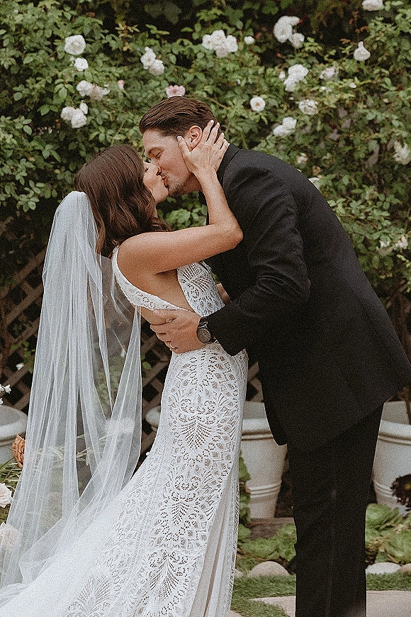 Wedding kiss portrait of bride and groom kissing, her lace wedding dress and long veil flowing against garden greenery and white roses