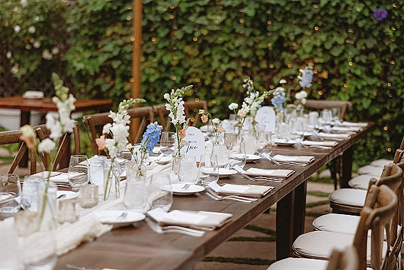 Reception tablescape on a long wedding table with wildflower bud vases, white napkins and plates, votive candles, and string lights by a greenery wall