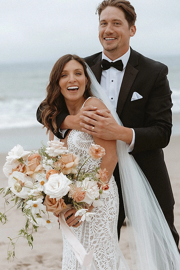 Couple portrait of bride and groom beach photo, groom hugging bride as she laughs, holding a peach rose bouquet with ocean backdrop