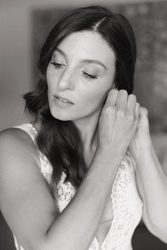 Bridal portrait close up of a bride with long lashes and side-swept waves in a lace wedding dress, against a softly blurred interior background