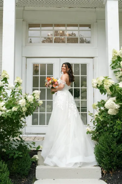 Bridal portrait of a bride holding bouquet in an off the shoulder lace wedding dress on porch steps by white French doors and hydrangeas