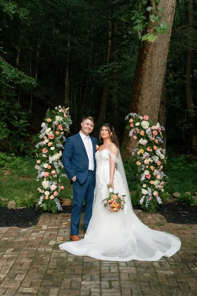 Couple portrait of bride and groom portrait with veil and bouquet beside floral pillars, set against forest trees on a brick patio