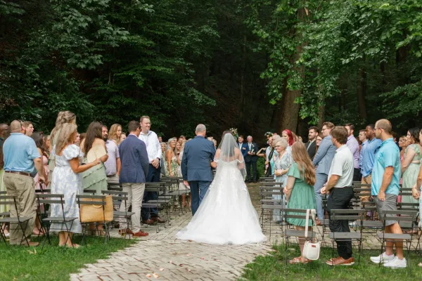 Wedding processional with bride walking down aisle beside her father, cathedral veil and gown train flowing on a brick walkway in a tree-lined garden