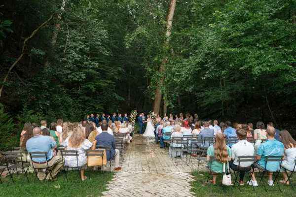 Outdoor wedding ceremony in a forest with guests seated along a brick aisle leading to a floral wedding arch and wedding party at the altar