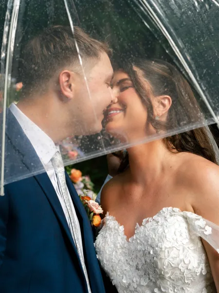 Wedding kiss portrait of bride and groom kiss under a clear umbrella, rain droplets and greenery behind, her strapless lace dress and his navy suit visible