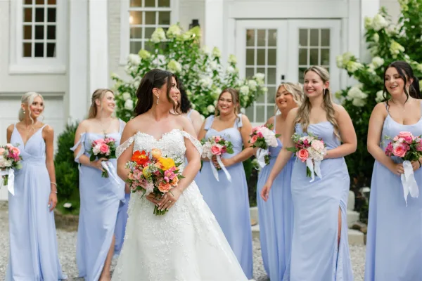 Bride with bridesmaids in light blue walking in front of a white house, holding colorful bouquets with ribbon tails and hydrangeas behind