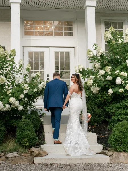 Couple portrait of bride and groom holding hands on white brick house steps, bride looking back in lace dress and cathedral veil with bouquet