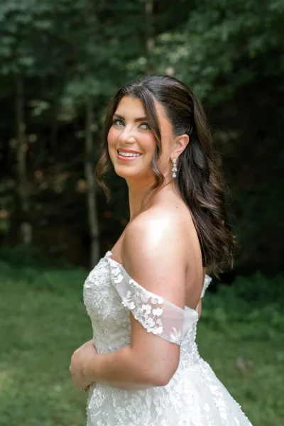 Bridal portrait of a bride looking over her shoulder in a lace off-the-shoulder gown with drop earrings, framed by forest greenery