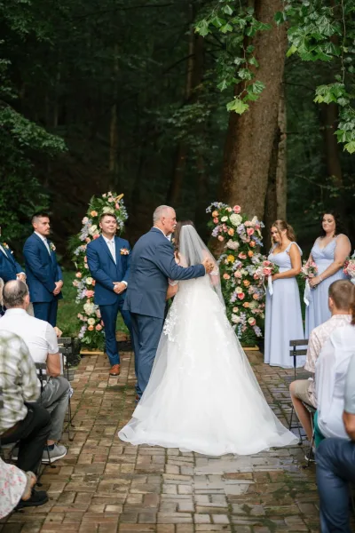 Processional with bride walking down aisle in cathedral veil and gown, flanked by wedding party on a brick walkway amid forest trees
