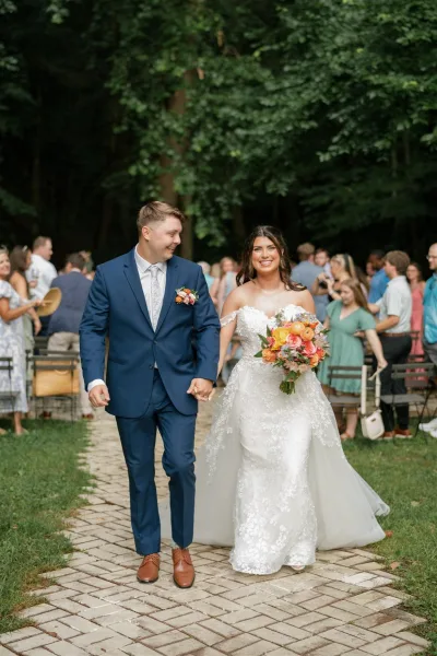 Wedding recessional as bride and groom walk the aisle holding hands, her colorful bouquet and lace dress flowing past guests on a tree-lined lawn