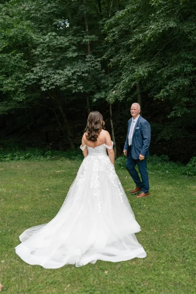 First look moment as bride reveal to groom in a lace off-the-shoulder gown with long train, approaching him in a forest clearing
