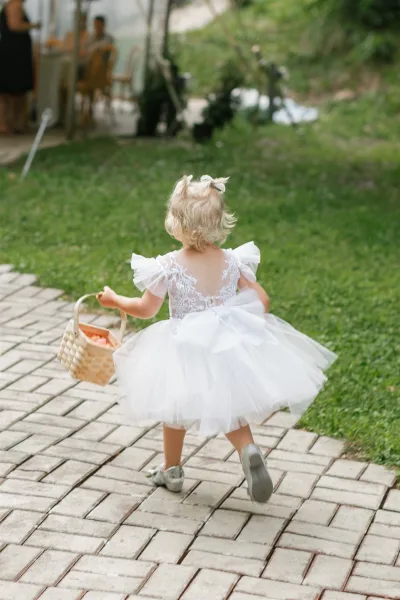 Flower girl in a flower girl dress with a hair bow carries a woven basket of petals on a brick walkway near an outdoor tent reception