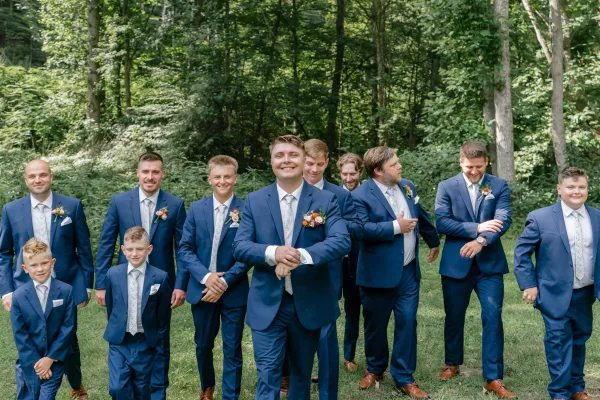 Groomsmen portrait of the groom with groomsmen in blue suits, ties and boutonnieres, walking on a grass lawn beneath woodland trees