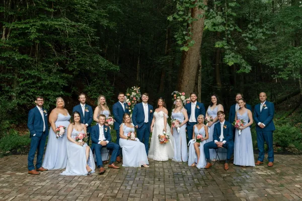 Wedding party portrait with bride, bridesmaids, groom and groomsmen in blue and light blue holding pink bouquets on a brick patio by forest trees