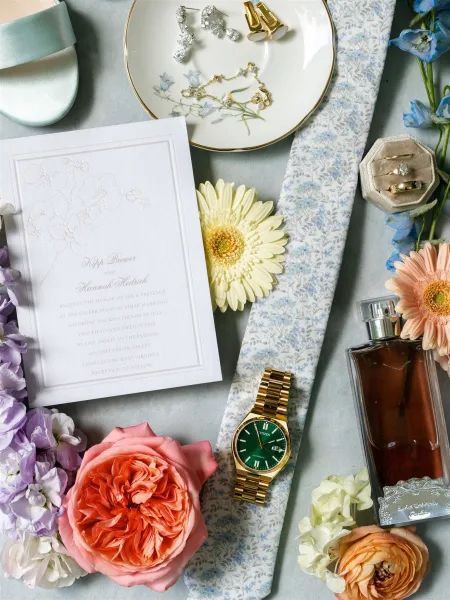 Invitation flat lay featuring a wedding invitation suite with ribbon, rings, jewelry, cufflinks, watch, cologne, and flowers on linen