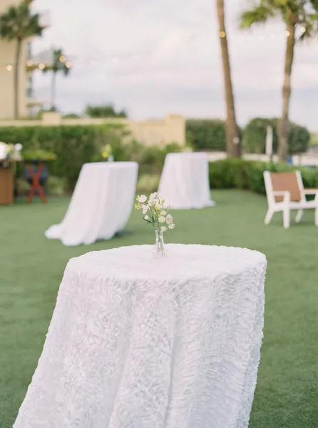 Cocktail table decor with a white textured tablecloth and bud vase of small white flowers, set on a lawn beneath string lights and palms