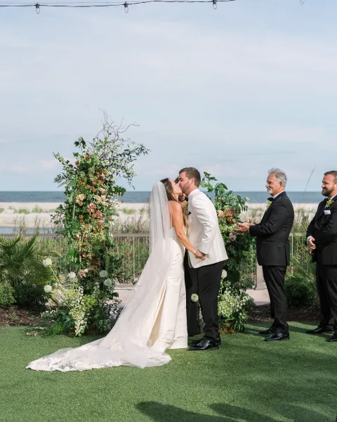 Wedding kiss at an oceanfront wedding ceremony under a floral arch with greenery and string lights, bride’s veil trailing behind groom in tuxedo