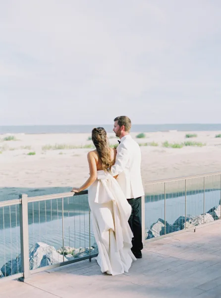 Couple portrait of bride and groom from behind, embracing at a balcony railing overlooking the ocean, her strapless dress flowing in sea breeze