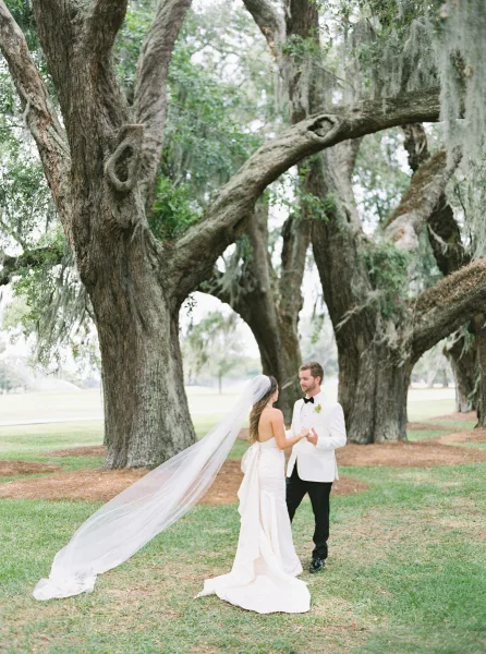Couple portrait of bride and groom holding hands under oak trees with Spanish moss, her long veil flowing beside his tuxedo boutonniere