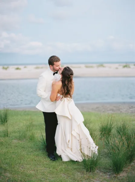 Wedding kiss portrait of bride and groom kissing on a beach, her strapless dress with bow back beside his white tuxedo, ocean dunes behind