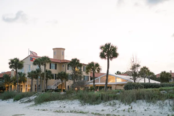 Wedding reception tent with string lights glowing on beach sand beside dune grass and palm trees under an open coastal sky