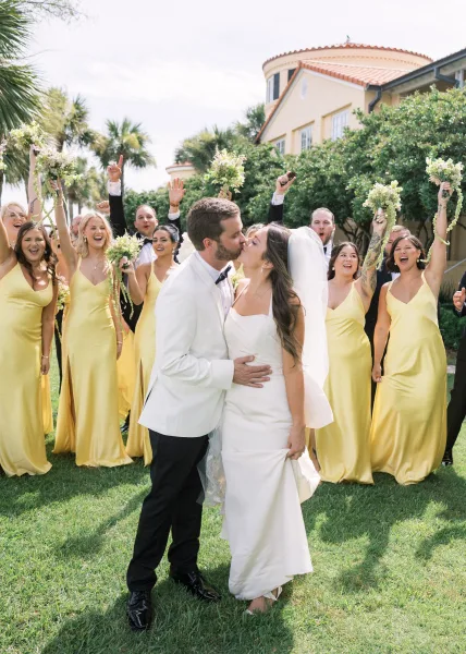 Wedding kiss portrait of the bride and groom kissing as the wedding party cheers around them on a palm-lined lawn by a stucco building