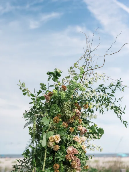 Wedding floral installation with greenery and blush roses on a ceremony floral pillar beside sand and grass under a blue oceanfront sky