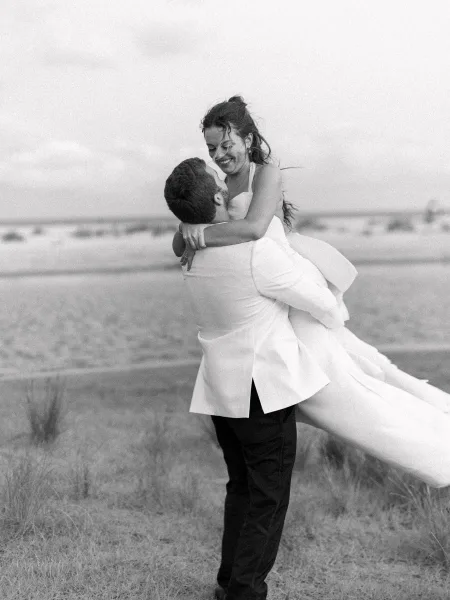 Couple portrait in a black and white wedding portrait as groom lifts laughing bride, her strapless dress train flowing by ocean sand dunes