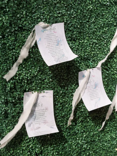 Wedding seating chart escort card display with calligraphy cards tied by white ribbon and string, hanging against a green boxwood wall