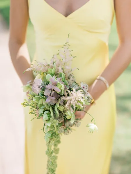 Wedding bouquet cascading with greenery and wildflowers, held with a bracelet in sunlit outdoor greenery background