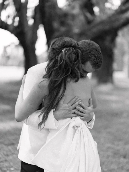 Wedding couple hug in a black-and-white outdoor portrait, groom in light suit embracing bride in backless dress on a tree-lined lawn