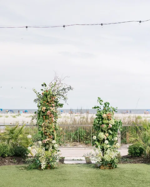 Wedding ceremony backdrop with floral ceremony pillars of roses, hydrangeas, and greenery on a beach lawn, with ocean and string lights behind