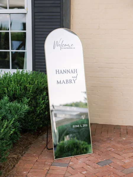 Wedding welcome sign on an arched mirror wedding welcome sign with a gold frame on a stand, set by a brick wall and shrubs outdoors