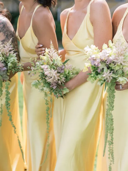 Bridesmaid bouquets held against yellow bridesmaid dresses, featuring greenery, trailing amaranthus and lavender flowers on an outdoor lawn