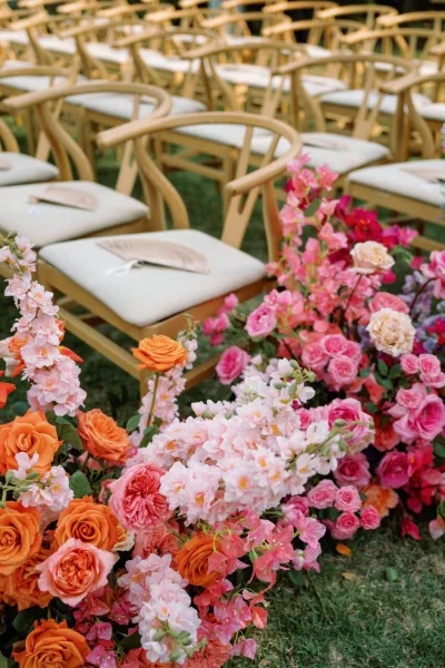 Ceremony aisle florals with bright pink and orange roses and sweet peas line a grass aisle beside wood chairs with hand fans