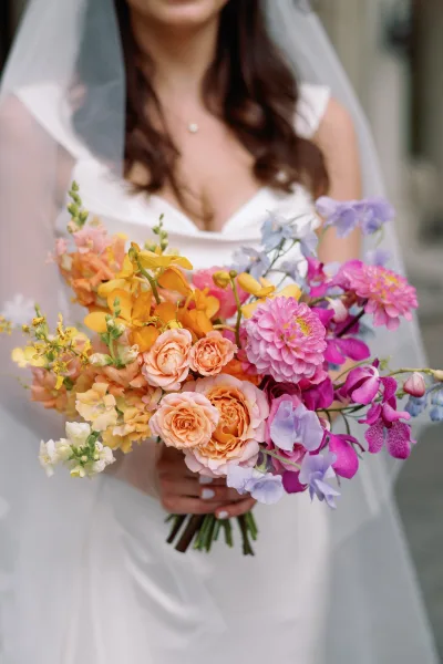 Bridal bouquet of roses, orchids, and sweet peas in a bright wedding bouquet, held against a bride’s white dress and veil outdoors.