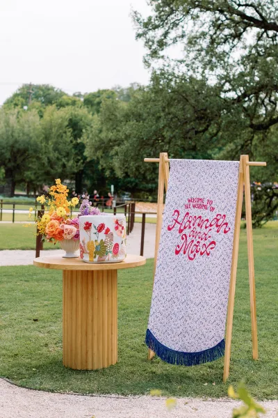 Wedding welcome sign on a wooden stand with fringe trim, beside a round table with single-tier cake and flowers on a park lawn