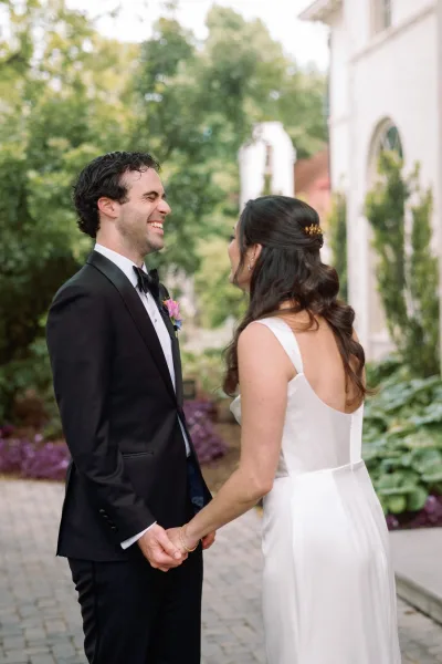 First look moment as bride in an open-back wedding dress meets groom in a black tuxedo on a cobblestone garden path with greenery