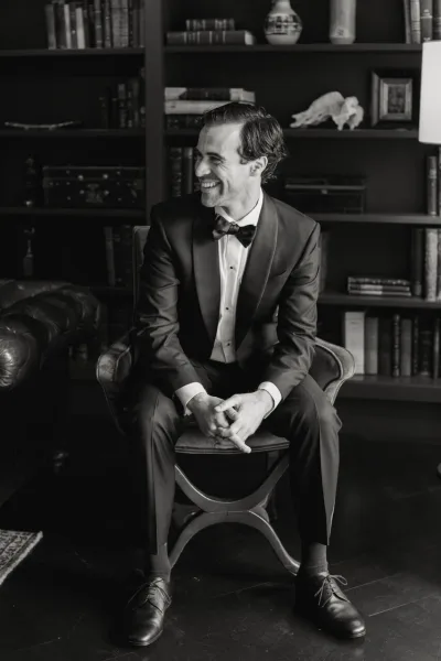 Groom portrait in a tuxedo with bow tie, seated in a lounge chair, smiling in a library room with bookshelves and a lamp